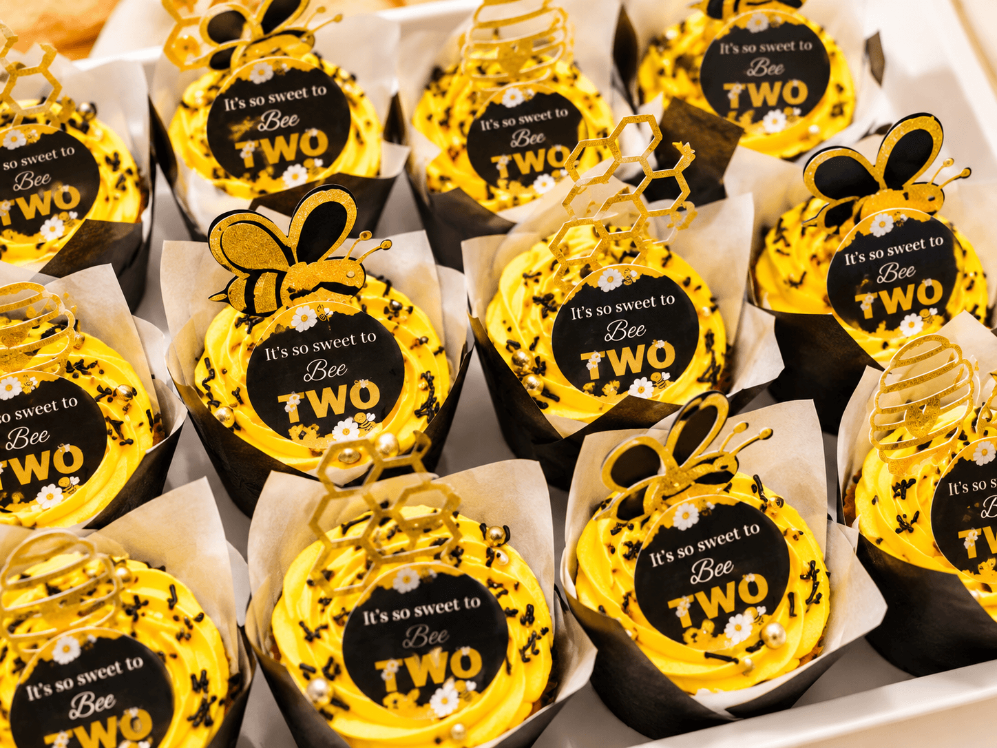 Yellow bee-themed cupcakes with "It's so sweet to Bee TWO" toppers and gold honeycomb picks
