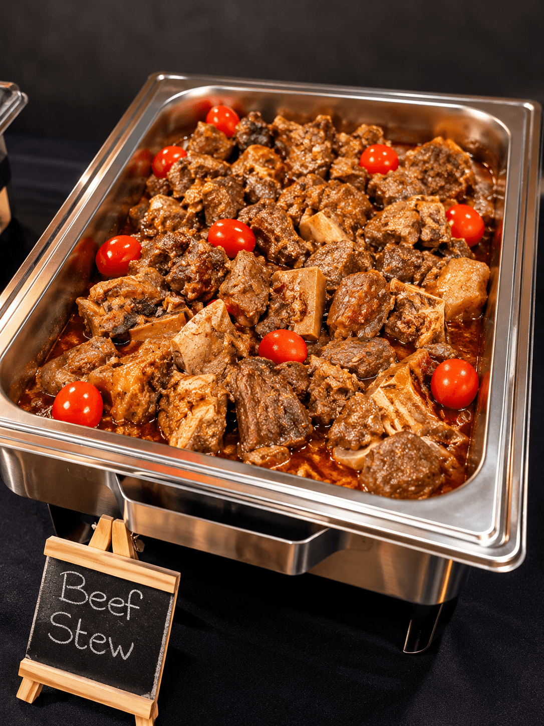 Chafing tray of slow-cooked beef stew with cherry tomatoes and a chalkboard label
