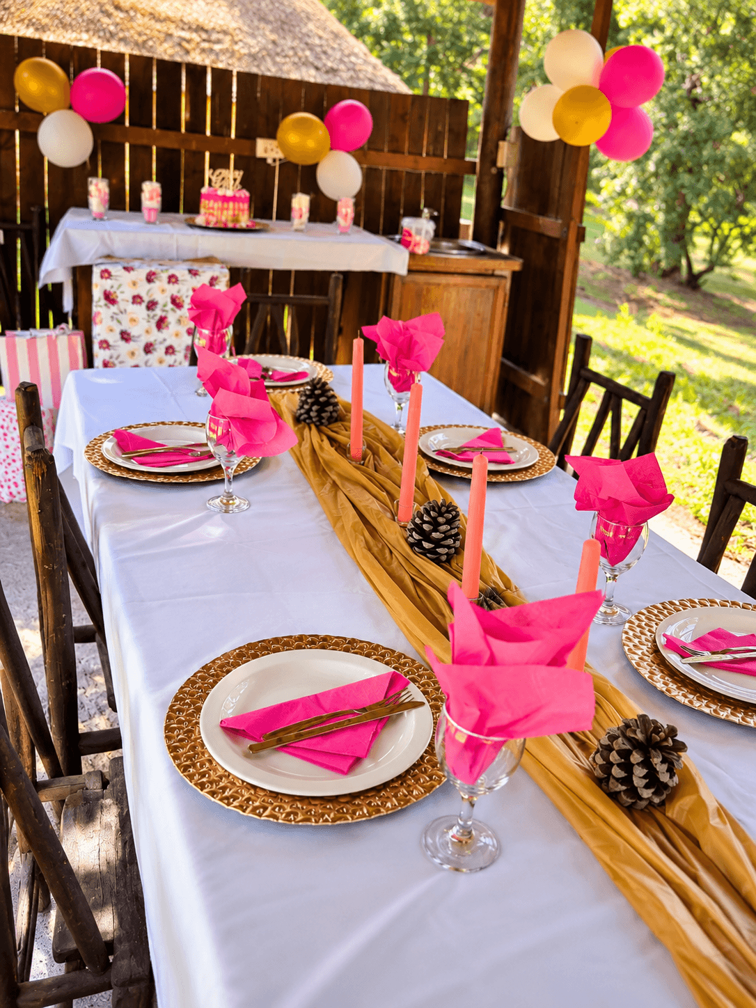 Garden party table with pink napkins, gold runner, coral candles and a celebration cake behind